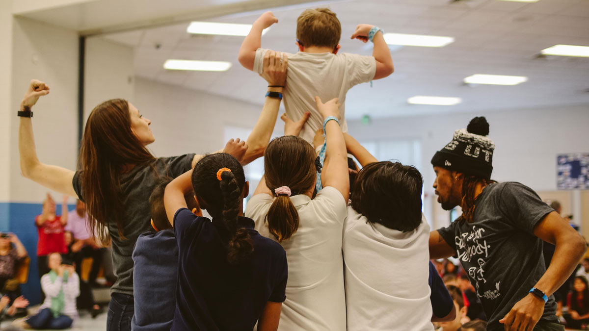 A female and a male dancer help four young children to lift a smaller boy as the female dancer and the smaller boy flex their biceps in a pyramid shape in front of an audience